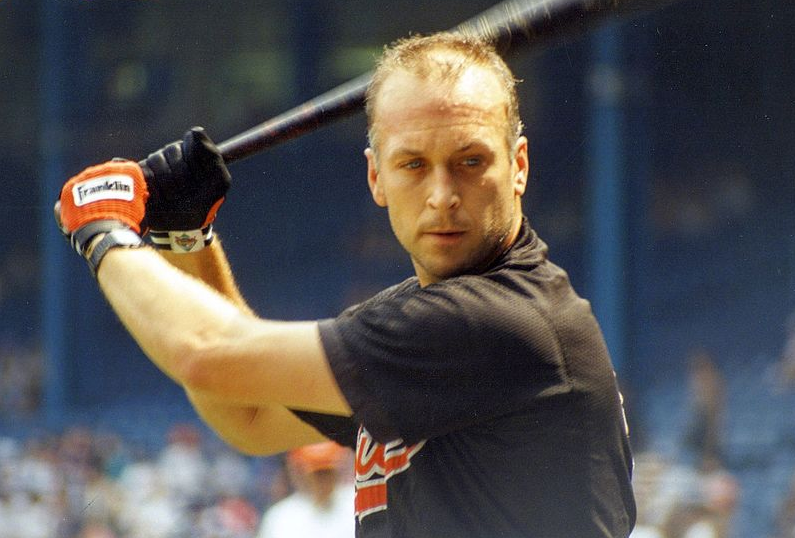 A baseball player wearing a black jersey and red Franklin batting gloves prepares to bat. He has a focused expression, holding the bat in a ready position. The background shows a stadium with spectators in the bleachers.