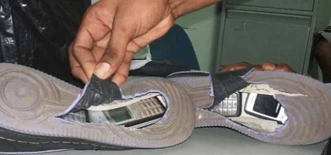 A pair of shoes with their inner soles partially removed reveals hidden mobile phones inside the soles. A person's hand is holding up the material of one shoe, showing the concealed phone.