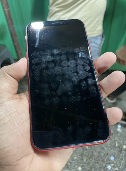 A hand holding a red smartphone with a touchscreen visibly covered in fingerprint smudges. The background is blurred but includes a person in a beige shirt and a green structure.