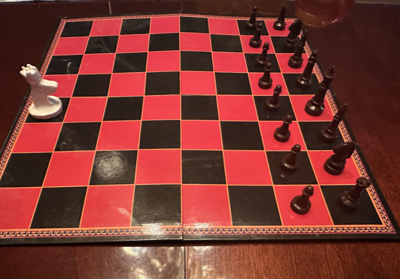 A chessboard with a black and red checkered pattern is set on a wooden table. One white knight is positioned on the left, while all black pieces (pawns, rooks, knights, bishops, queen, and king) are arranged on the right side in their starting positions.