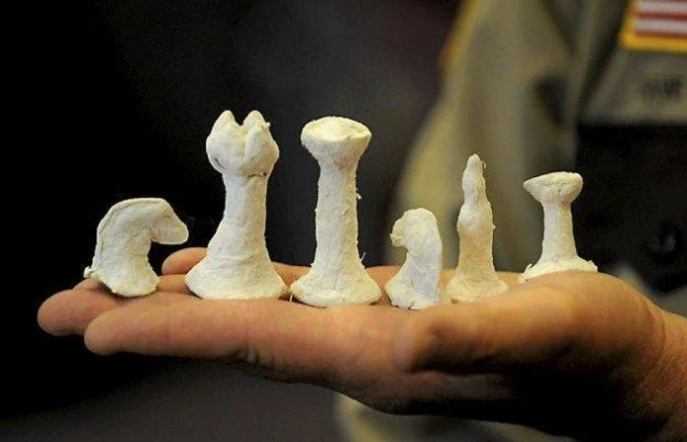 A person's hand holding six roughly sculpted, white clay chess pieces of varying sizes and shapes. The background is slightly blurred, focusing attention on the pieces.