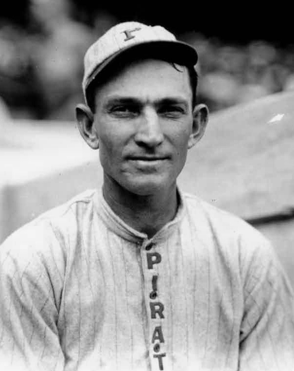 Black and white photo of a baseball player from the early 20th century. He is wearing a uniform with vertical stripes and a cap. The word "PIRATES" runs down the front of his jersey. The background is blurred, focusing attention on the player.