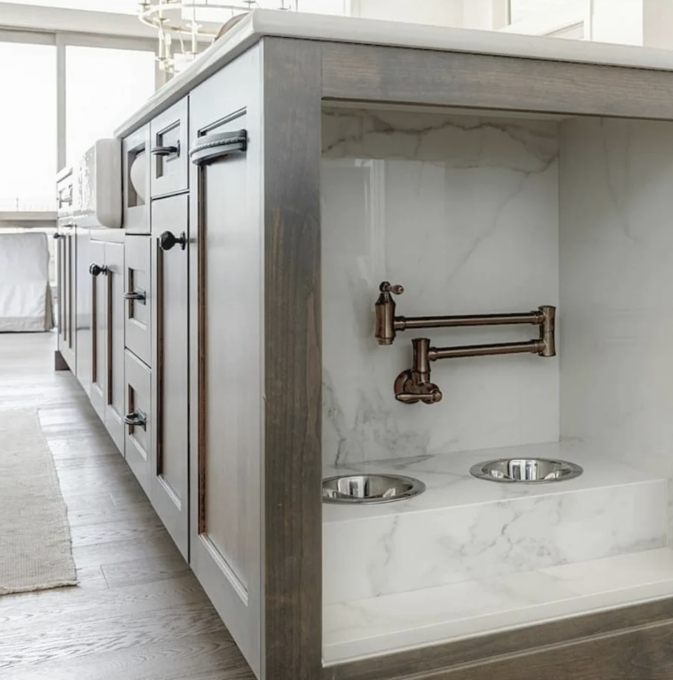 A modern kitchen island featuring dark cabinetry and a marble backsplash. The island includes a built-in pet feeding station with two stainless steel bowls and a wall-mounted faucet, all set within a sleek, dedicated alcove. The floor is wooden and partially covered by a rug.