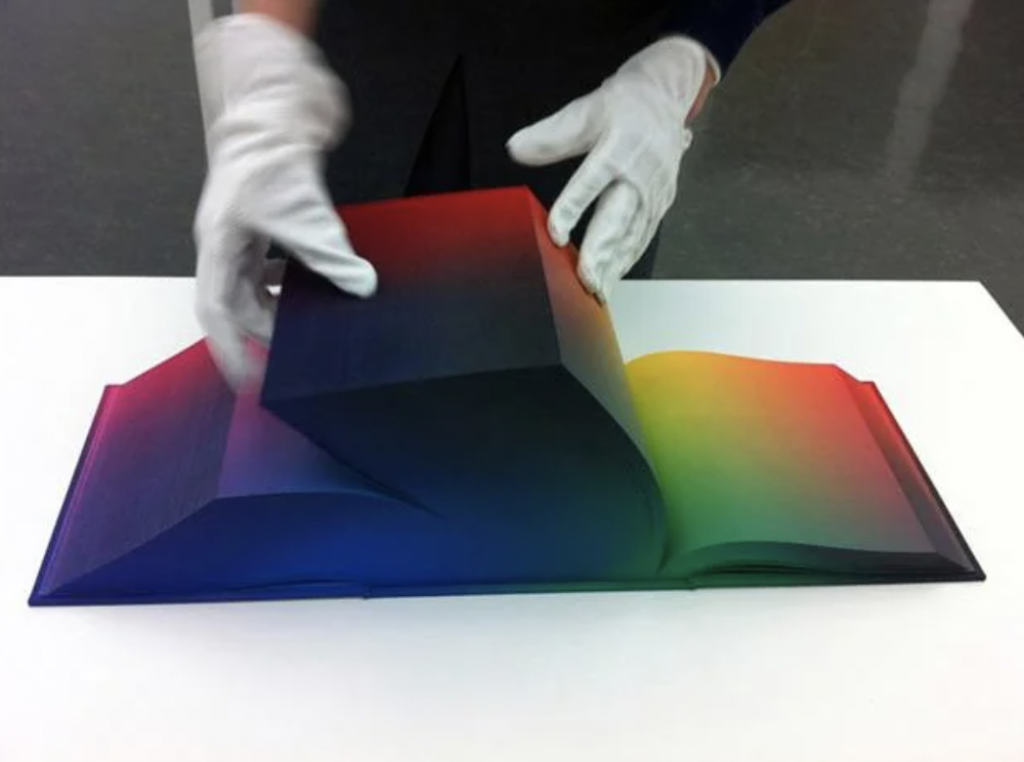 A person wearing white gloves is handling an open, thick book placed on a white surface. The book's pages have a gradient color transition, moving from blue to green to red, creating a visually striking effect.