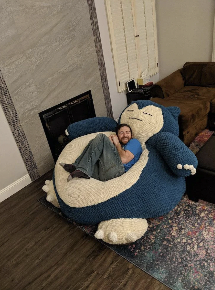 A man with a beard happily lies on a large, plush Snorlax bean bag chair in a living room. The bean bag, shaped like the popular Pokémon character Snorlax, is blue and white and takes up a significant portion of the space. The room has a cozy, casual feel.