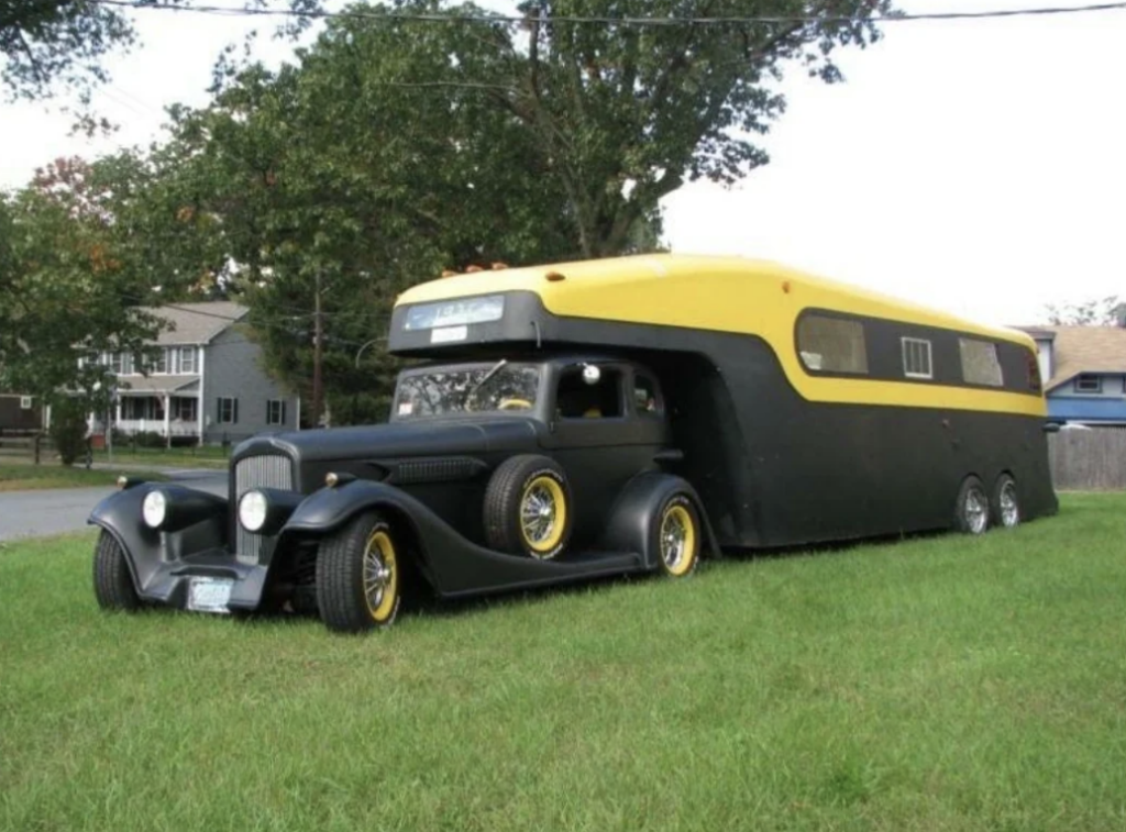 A vintage black and yellow hot rod car is modified into a unique RV on a grassy lawn. The RV has the chassis of the car in front and extends into a modern-style camper at the rear, with large windows along the side and three axles with chrome wheels.