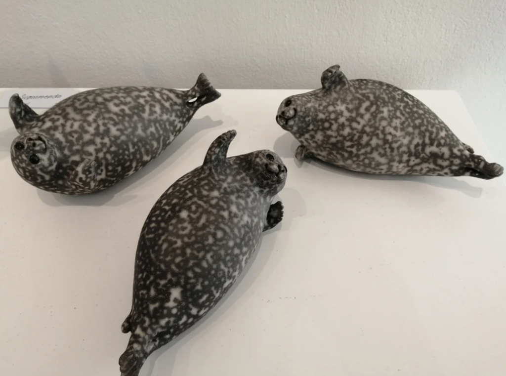 Three black-and-white ceramic sculptures of seals rest on a white surface. The seals have spotted patterns and are in various relaxed poses, seemingly lounging. The background is a plain, light-colored wall.