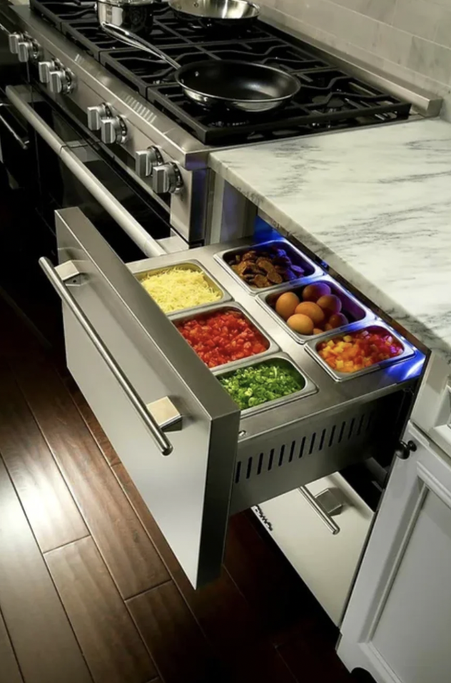 A modern kitchen with a stove and marble countertop. An open drawer beneath the countertop reveals compartments with various ingredients including grated cheese, diced tomatoes, chopped green onions, sliced mushrooms, whole eggs, and chopped bell peppers.