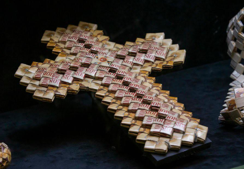 A cross made from tightly woven strips of materials, including visible "10$" labels, lies on a dark surface. The design is intricate, displaying a mix of light and dark hues, giving it a textured appearance.