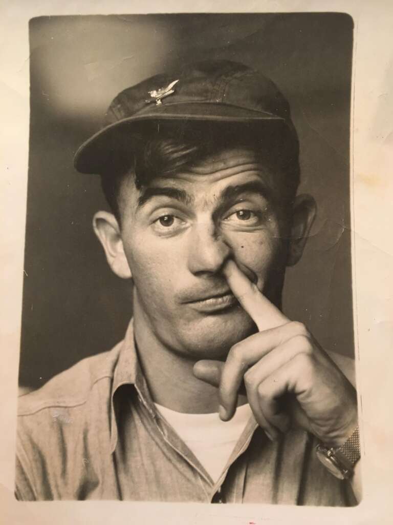 A black-and-white photo of a man wearing a baseball cap and a buttoned-up shirt. He has a playful expression on his face and is sticking his index finger up his nose. He is looking directly at the camera.