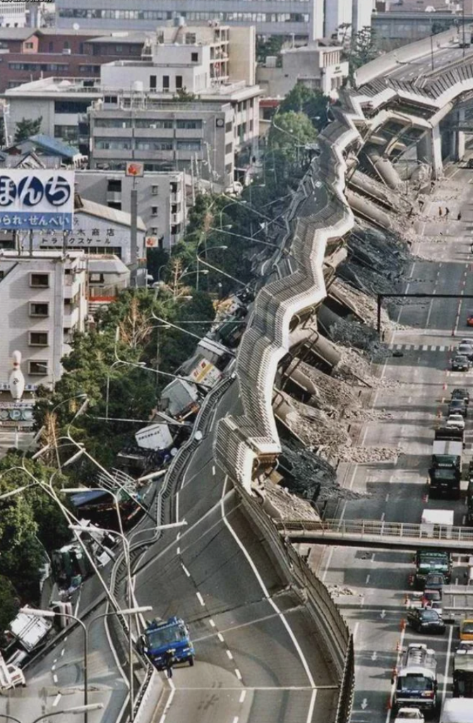 A damaged elevated highway with cars trapped on it after it collapsed during an earthquake in an urban area. Surrounding buildings and infrastructure are also affected, with debris and destruction evident. Emergency vehicles and responders are present on the scene.