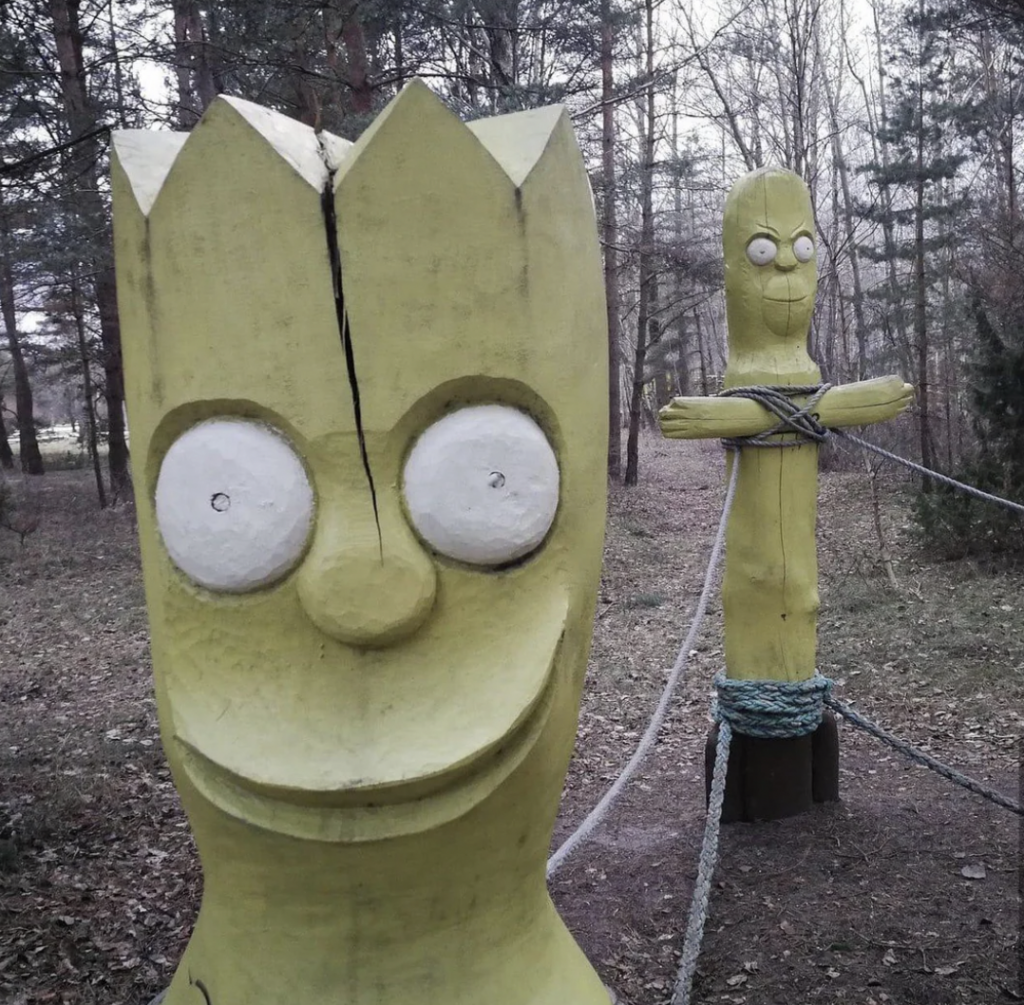 Two yellow wooden sculptures resembling cartoon characters stand in a forest. The foreground sculpture shows a face with wide white eyes and a cracked head. The background sculpture is bound with ropes around its neck and arms, creating a cross-like shape.