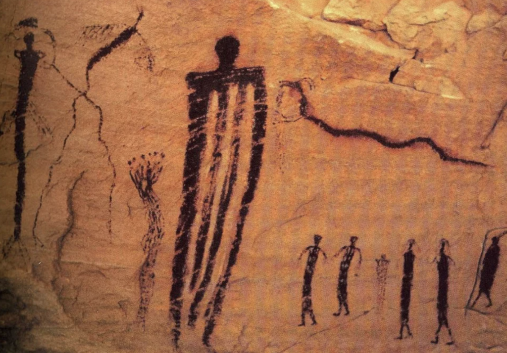 A photograph of ancient rock art depicting several humanoid figures. The figures include one tall dark silhouette with vertical stripes, several smaller solid figures, and a few faint, less distinct humanoid shapes. The background is a reddish-brown rock surface.