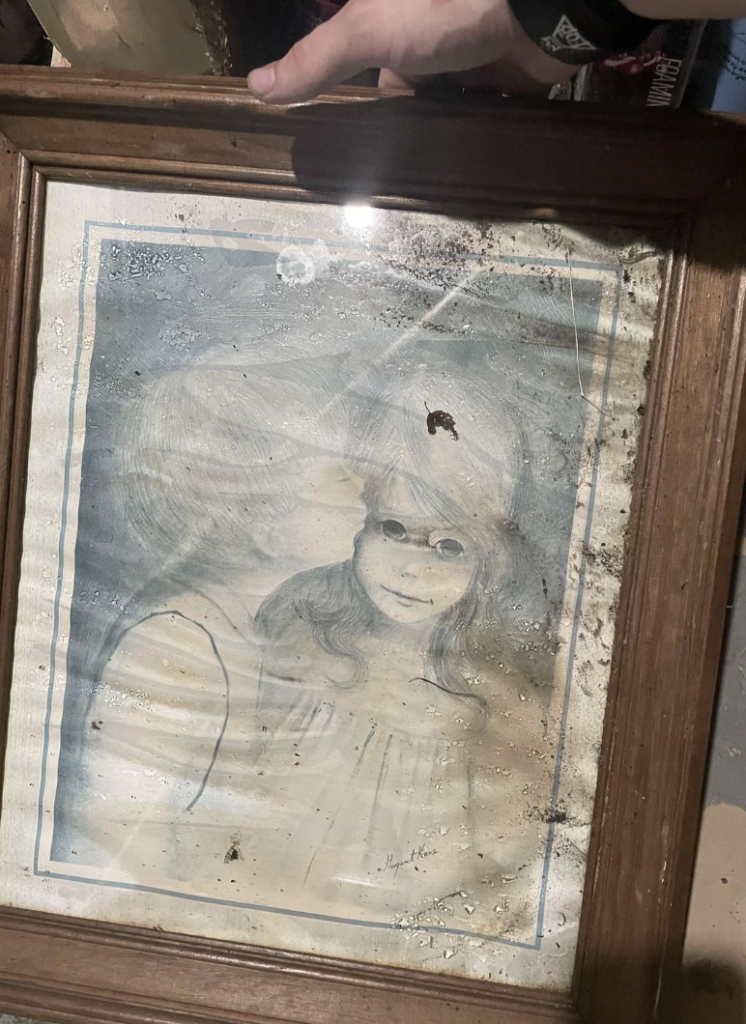 A person holds an old, weathered, framed painting of a child being kissed on the forehead by another person. The painting is faded and stained, indicating damage over time. The frame is wooden and shows signs of age.