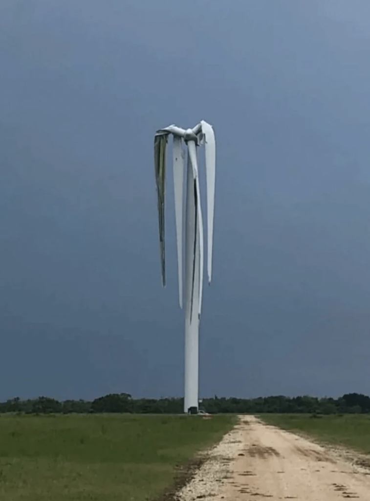 A damaged wind turbine with its blades visibly bent and hanging down is set against a dark, stormy sky. The turbine stands tall at the end of a dirt path, surrounded by a field with a line of trees in the distance.