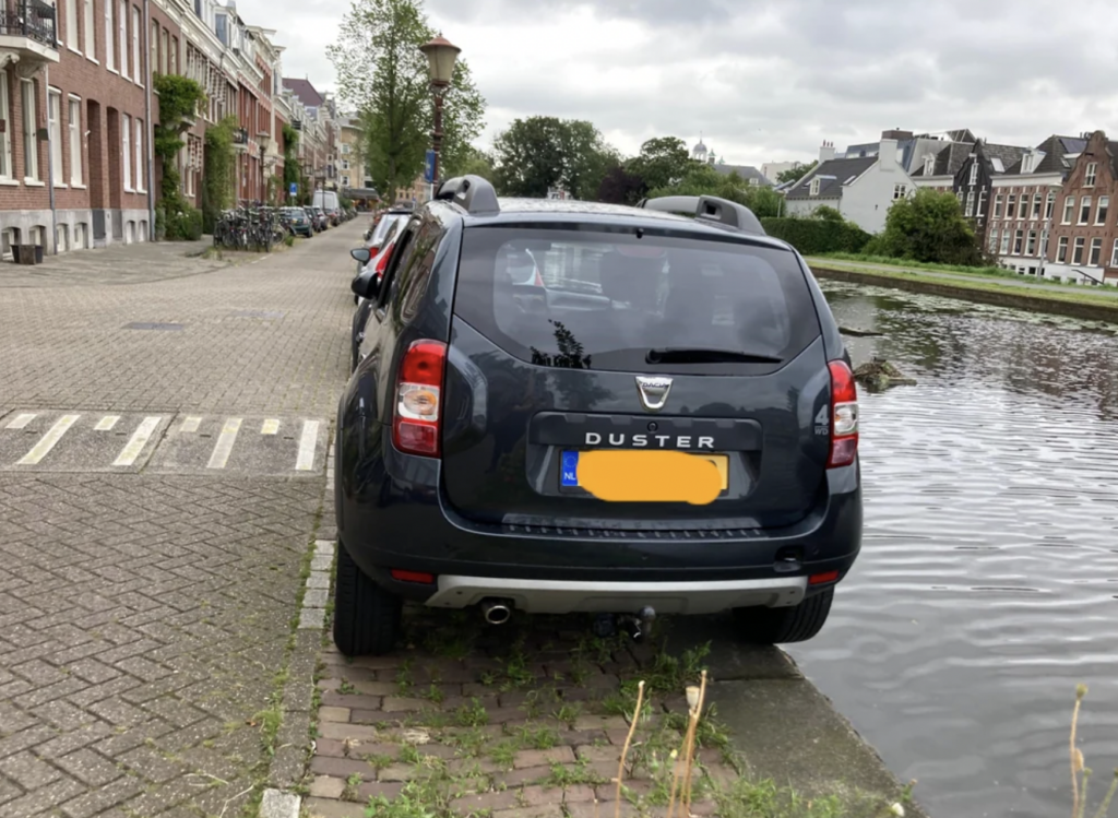 A black Dacia Duster is parked partially on a sidewalk and grass next to a canal with water on the right side. The background features buildings and trees under an overcast sky. The car's license plate is partially obscured for privacy.