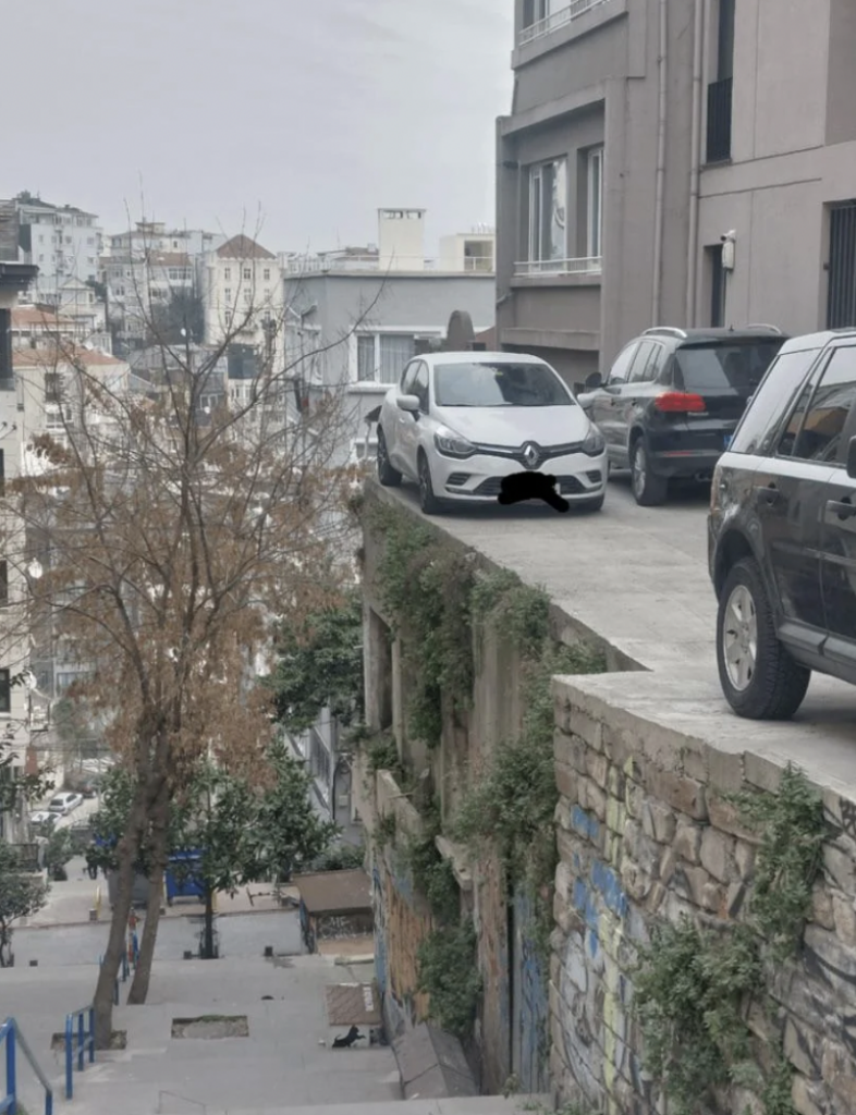 A white car is parked precariously on the edge of a high wall, with a significant drop to the street below. The car is alongside a black vehicle, both near a building. In the background, there is an urban landscape with various buildings and a tree with bare branches.