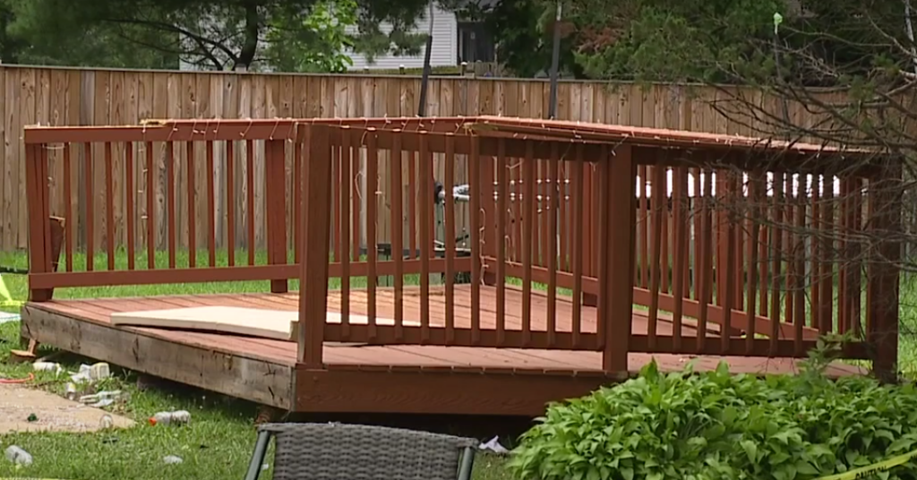 A raised wooden deck with a simple railing stands in a backyard with a grassy lawn. The deck is stained a reddish-brown color and is surrounded by a wooden fence. Some garden furniture and a few scattered items can be seen around the deck.