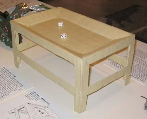 A small table model, possibly made from folded paper, displayed on a surface. Two six-sided dice, each showing two dots, are placed on top of the table. There are text documents and other items partially visible around the model.