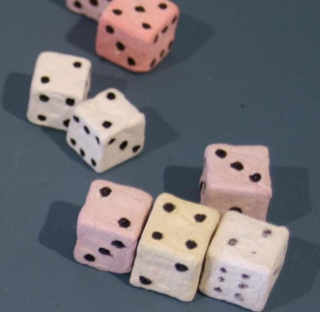 Soft pastel-colored decorative dice with black dots are arranged randomly on a dark blue surface. There are six dice in total, with colors including pink, white, and light yellow, each showing various numbers on their faces.