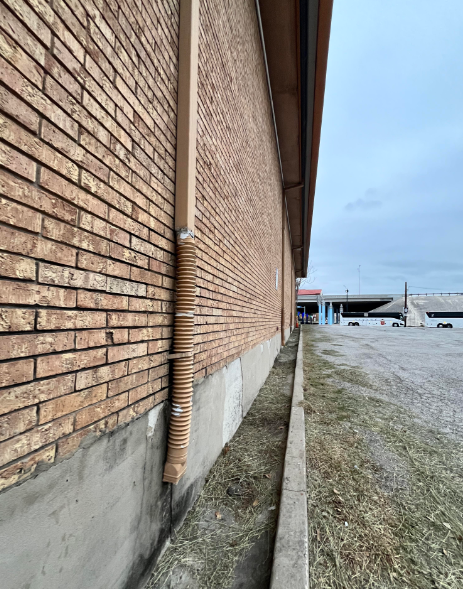 A long, brown brick building with a tan downspout runs parallel to a narrow gravel path. The sky is overcast. A few cars are visible in the distance near an overhead structure. The image is taken at an angle, emphasizing the length of the building.