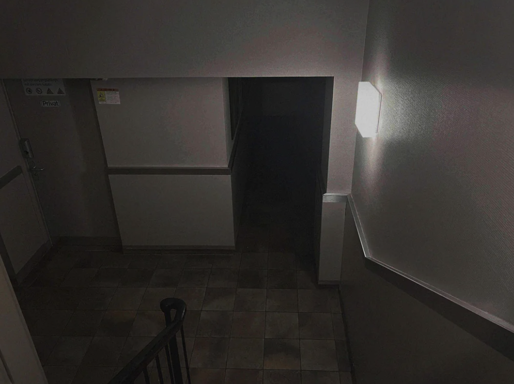 A dimly lit, empty hallway with tiled floor and stair railing on the left. The hallway leads to a dark doorway. A bright wall light is on the right, casting shadows on the walls. A sign on the left wall points to a private area.