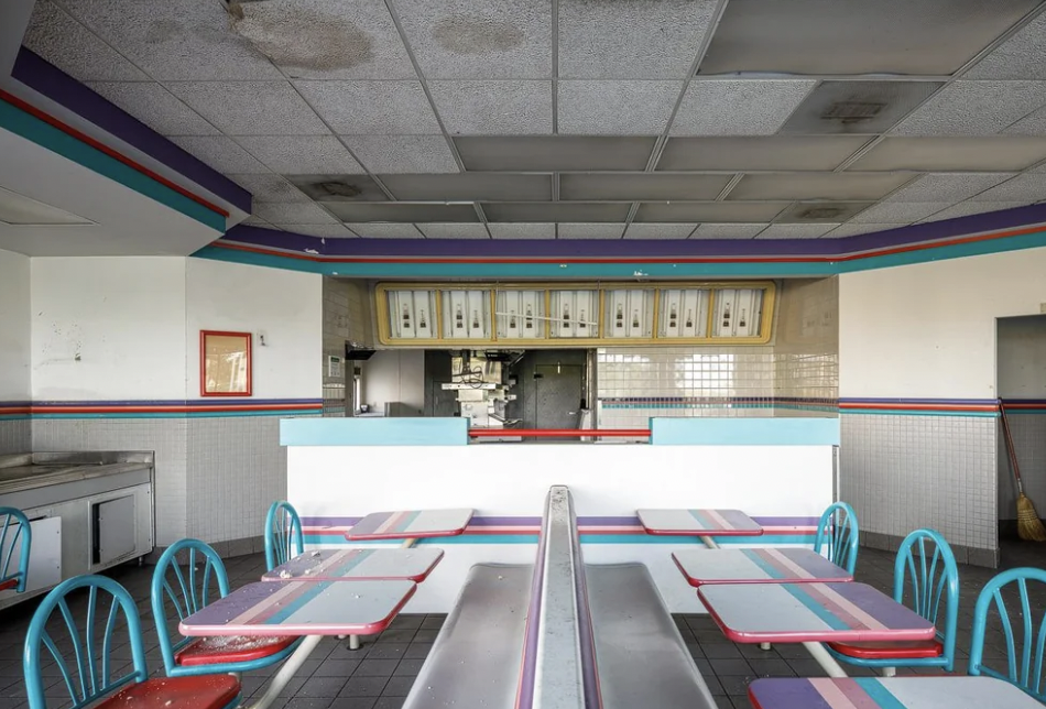 An abandoned fast-food restaurant with discolored ceiling tiles, outdated colorful furniture, and tables with debris. The booth seating features a faded teal and pink color scheme. The counter area in the background shows an empty kitchen space.