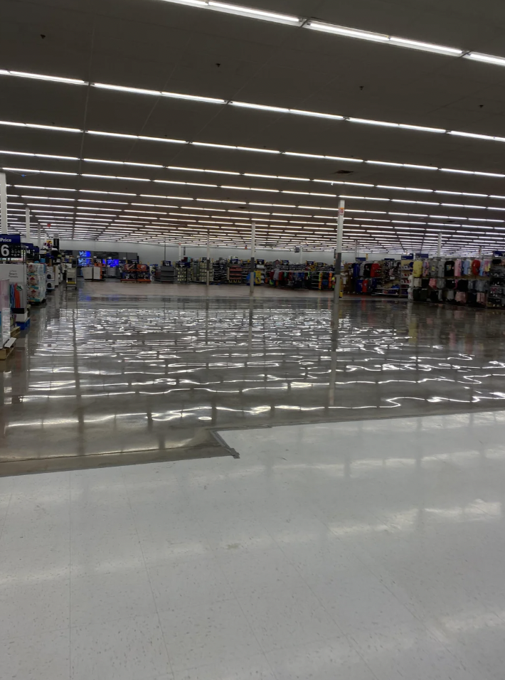 A large, brightly lit retail store with aisles of products on either side. The highly polished floor reflects the overhead fluorescent lights, creating a mirror-like effect. Clothing and other items are neatly arranged on racks and shelves throughout the store.