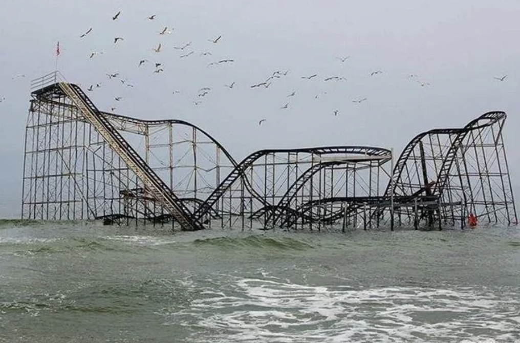 Partially submerged roller coaster in the ocean, surrounded by waves and flying seagulls against a cloudy sky. The structure is rusted and appears abandoned.