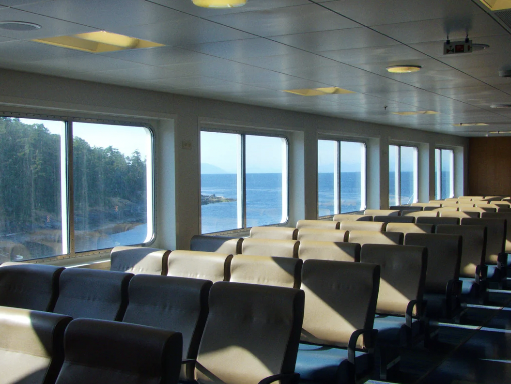 Interior of a ferry boat with rows of empty seats facing large windows. The windows offer a scenic view of the ocean, distant mountains, and a forested shoreline. Sunlight filters in, casting shadows on the seats and floor.