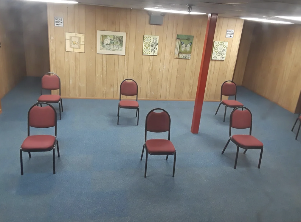 A room with wooden paneling, blue carpet, and six spaced-out red chairs facing a wall. The wall has various framed artworks displayed, and a red pillar stands centrally among the chairs. A "Keep Room Neat" sign is posted on the wall.