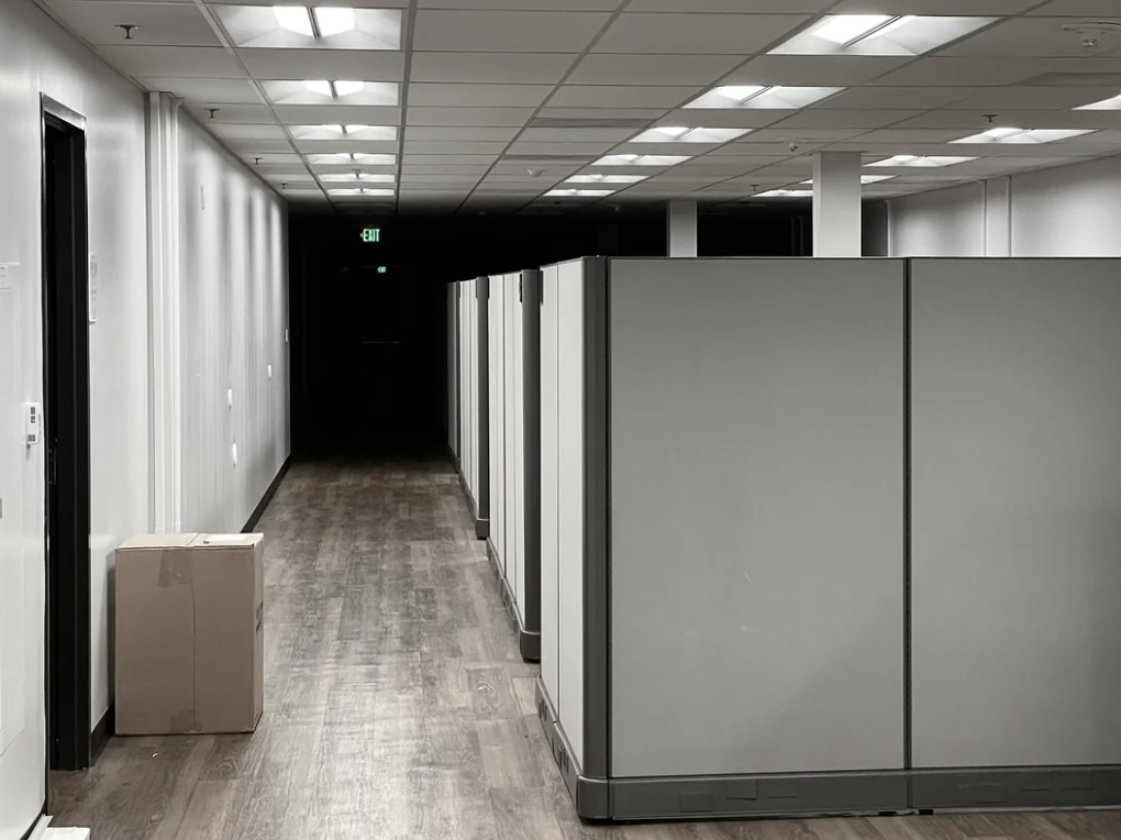 A dimly lit office hallway with modern wooden flooring and rows of tall, grey cubicle walls on the right. A cardboard box is placed on the floor near the entrance on the left. The corridor leads to a dark area in the distance.