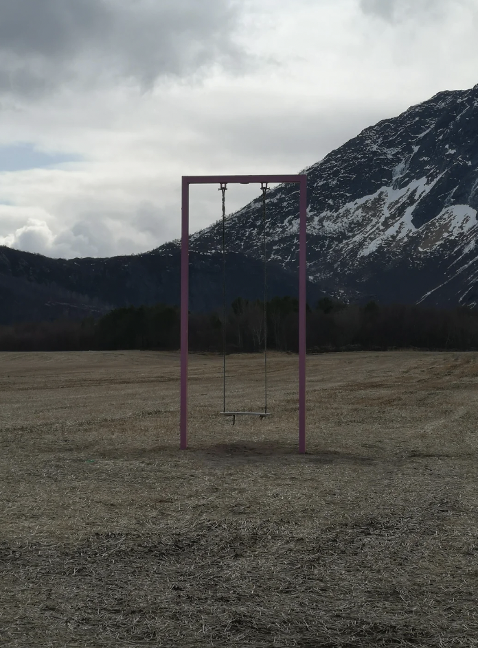 A solitary swing with a purple metal frame stands in a barren field. Snow-capped mountains and a cloudy sky create a dramatic backdrop. Trees are visible at a distance. The scene evokes isolation and tranquility.