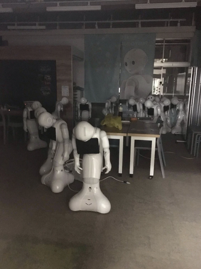 A dimly lit room filled with a group of humanoid robots. The robots are white and have smooth, rounded forms with digital screens on their chests. They are positioned in various postures, with some leaning forward and others standing upright.