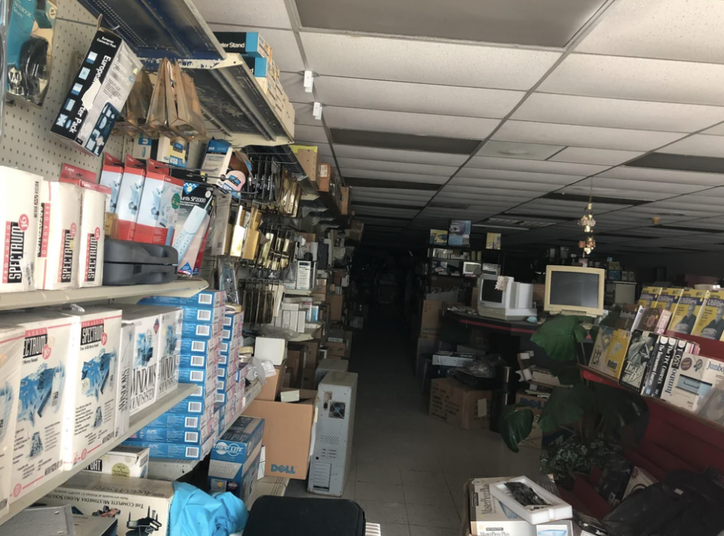 A dimly lit electronics store aisle filled with various boxed items, cables, and components on shelves. Old computer equipment, monitors, and other electronic devices are scattered around. The store appears cluttered, with limited walking space.