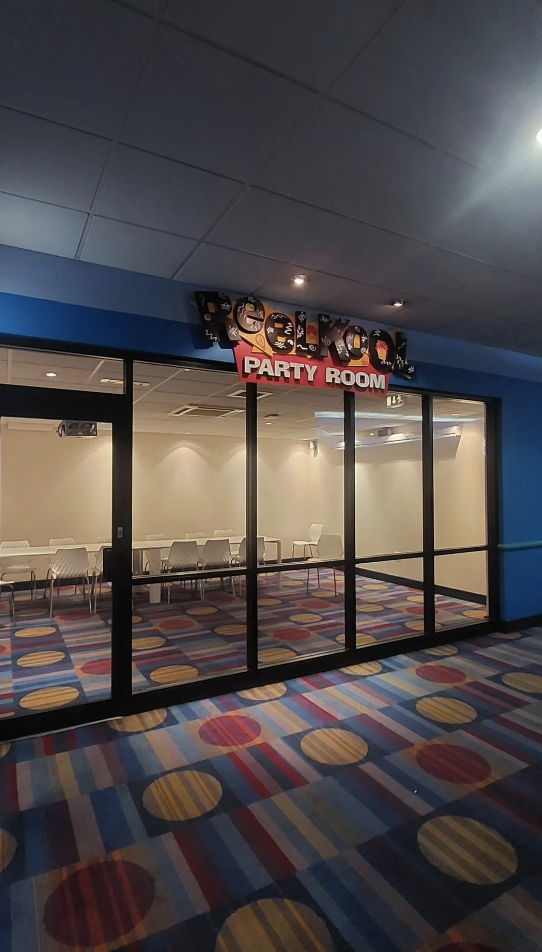 A glass-walled party room with a sign above the entrance reading "Party Room" in red letters. The room has white chairs arranged around tables and colorful, patterned carpet. The exterior hallway also features a multi-colored, patterned carpet.