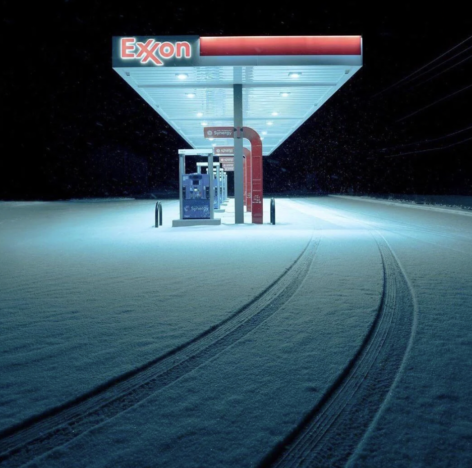 A deserted Exxon gas station at night with a snowfall-covered ground. Two sets of tire tracks lead up to the pumps, which are illuminated under the station's canopy. The scene is calm and quiet, with the dark night sky in the background.