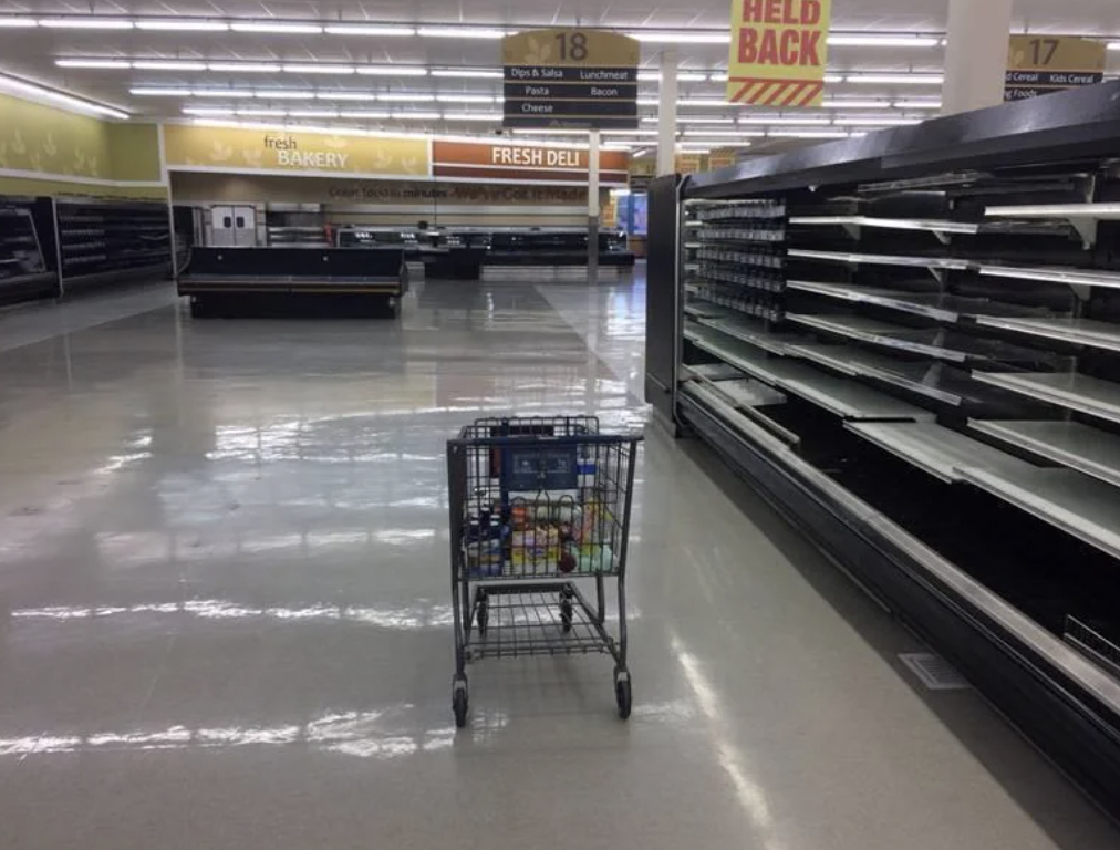 An empty grocery store aisle with only one shopping cart containing various items. The shelves are largely empty, and the lighting creates a somewhat deserted atmosphere. Signs for the bakery and deli sections are visible in the background.