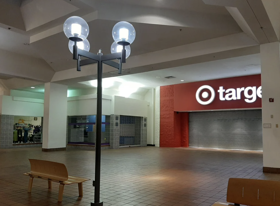 An empty shopping mall corridor featuring a streetlamp-style light fixture, two wooden benches, and a closed Target store with its metal shutter down. The Target sign is illuminated, and the atmosphere appears quiet and deserted.