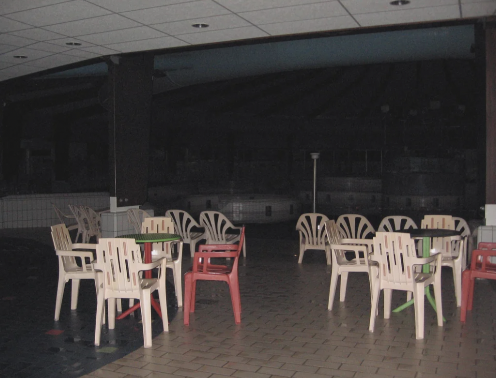 An indoor setting with white, pink, and green plastic chairs around tables on a tiled floor. The area appears dimly lit and empty, and large windows or openings in the background show an even darker space. The ceiling has recessed lighting.