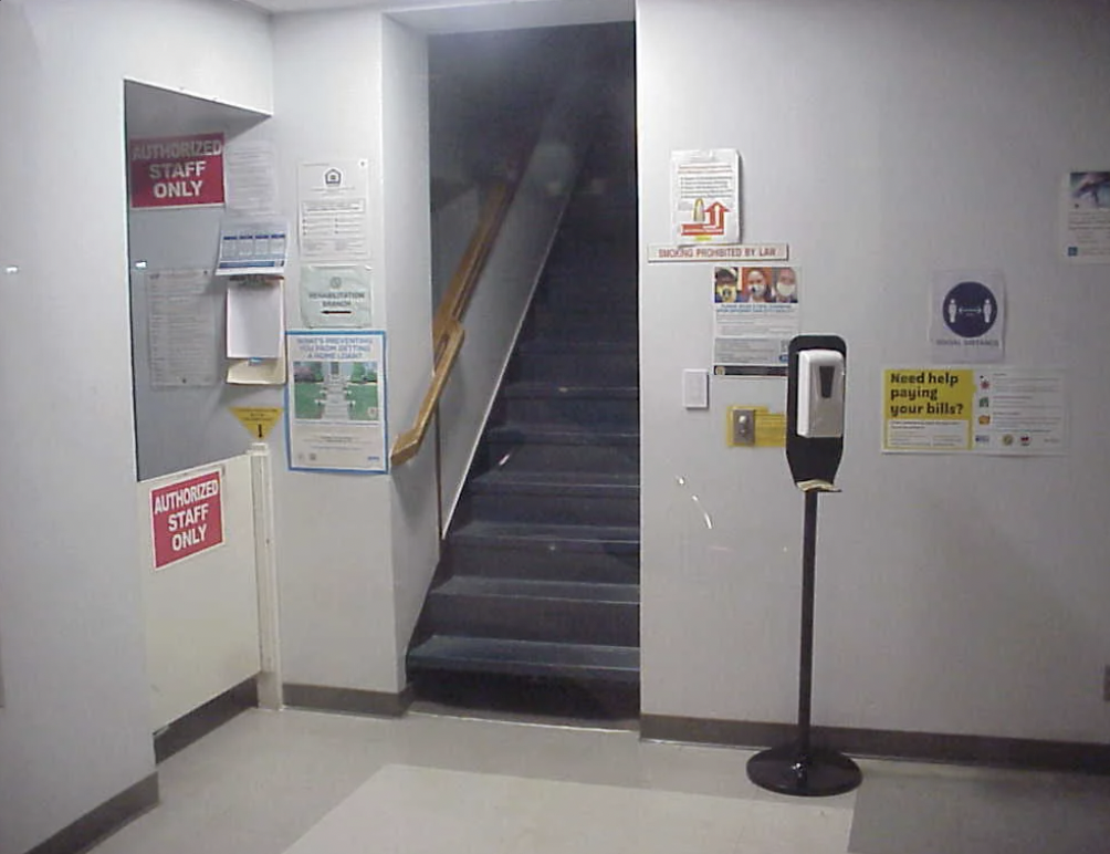 A stairway marked "Authorized Staff Only" is at the center. Surrounding walls display various signs including health and safety information and a notice about assistance with bills. A hand sanitizer station is to the right of the stairs.