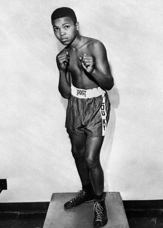 A young boy stands on a platform in a boxing pose with his fists up. He is wearing boxing gloves, shorts, and boxing shoes. The shorts have "LOUISVILLE KY" written on the side. The background is plain.