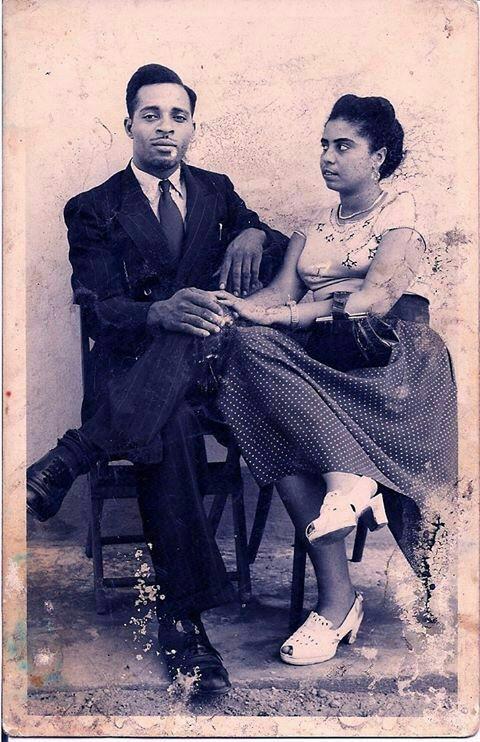 A vintage black-and-white photograph shows a man and a woman sitting closely together. The man, dressed in a suit, has one arm around the woman, who is wearing a polka-dot dress. They both have serious expressions and are seated on wooden chairs against a plain background.