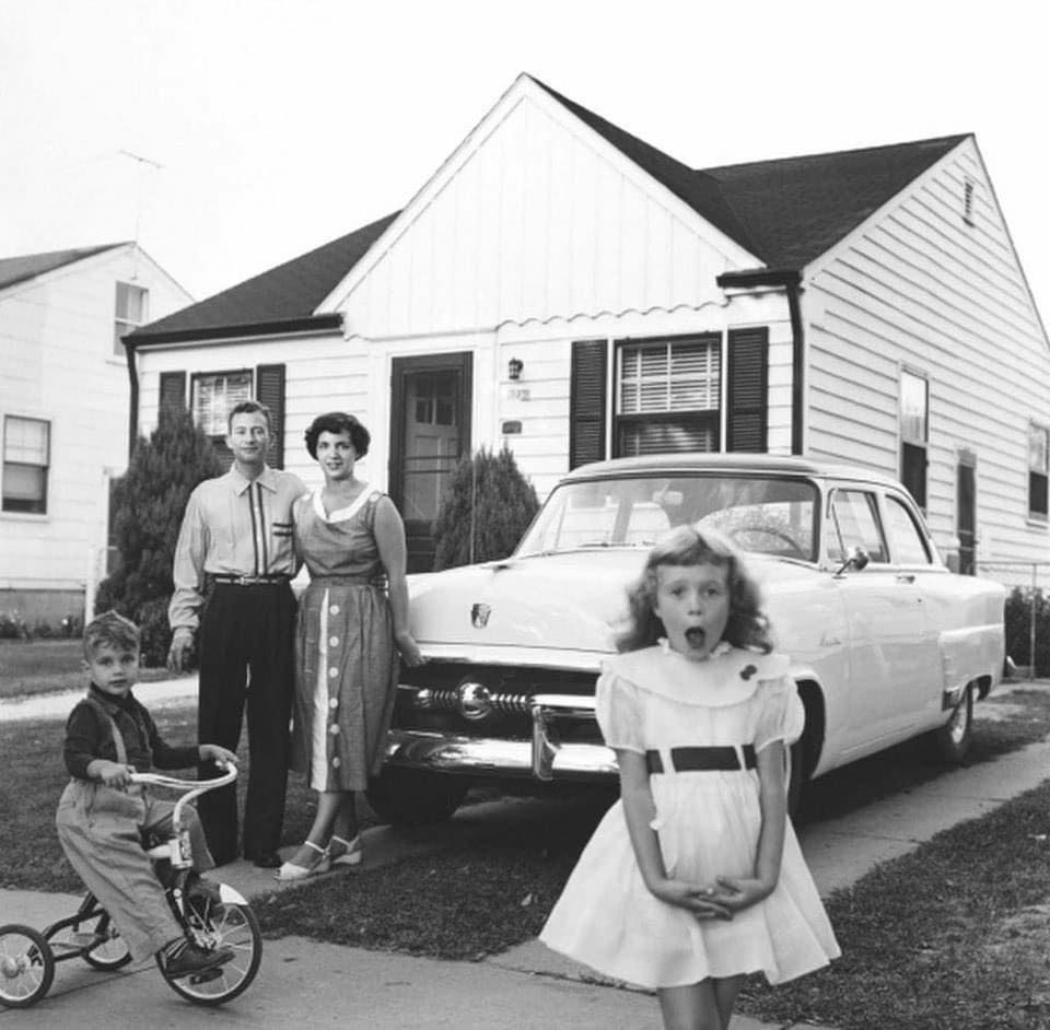 A 1950s family stands in front of their house and parked car. The father, mother, and son on a tricycle pose near the curb, while the daughter stands in the foreground with an expressive look on her face. The house features white siding and a triangular roof.