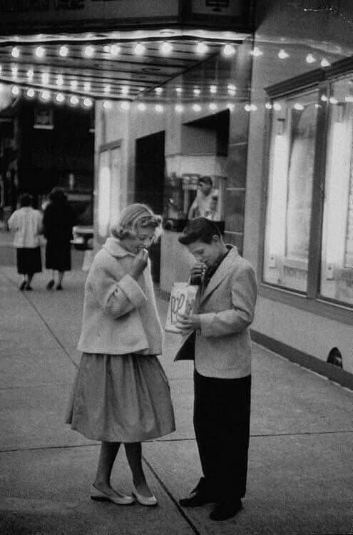 Black and white photo of a young boy and girl under marquee lights outside a theater. The boy is drinking from a straw while holding a popcorn bag, and the girl is looking down with her hand near her face. Other people can be seen in the background.