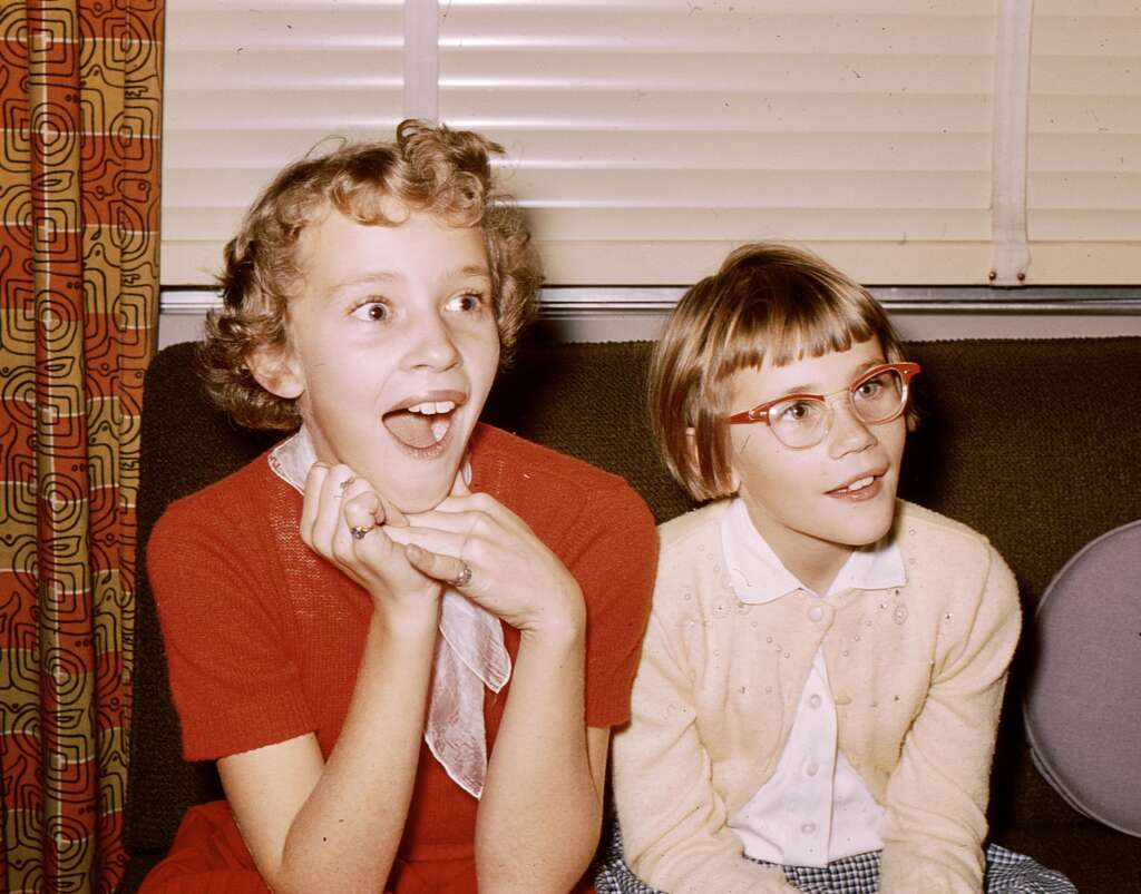 Two young girls sit side by side on a couch. The girl on the left, with curly blonde hair, is wearing a red dress and looks surprised with her hands under her chin. The girl on the right, with straight brown hair and red glasses, is wearing a white cardigan and is smiling.