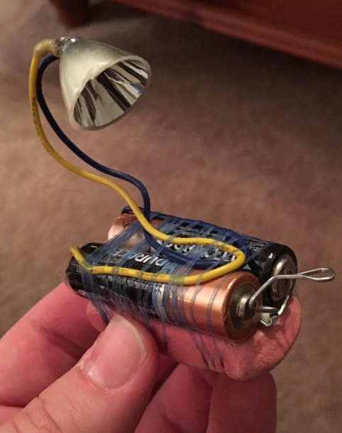 A handheld makeshift device features two AA batteries connected in series with insulating tape, a small lightbulb at the end of two yellow and blue wires, and a paperclip securing the circuit. The device is being held between two fingers.