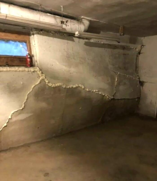 A basement with a cracked and bulging concrete wall, showing signs of structural damage. The crack has been filled with an unidentified material. There is a small window near the ceiling and some pipes running along the wall. A red extinguisher sits on the window ledge.