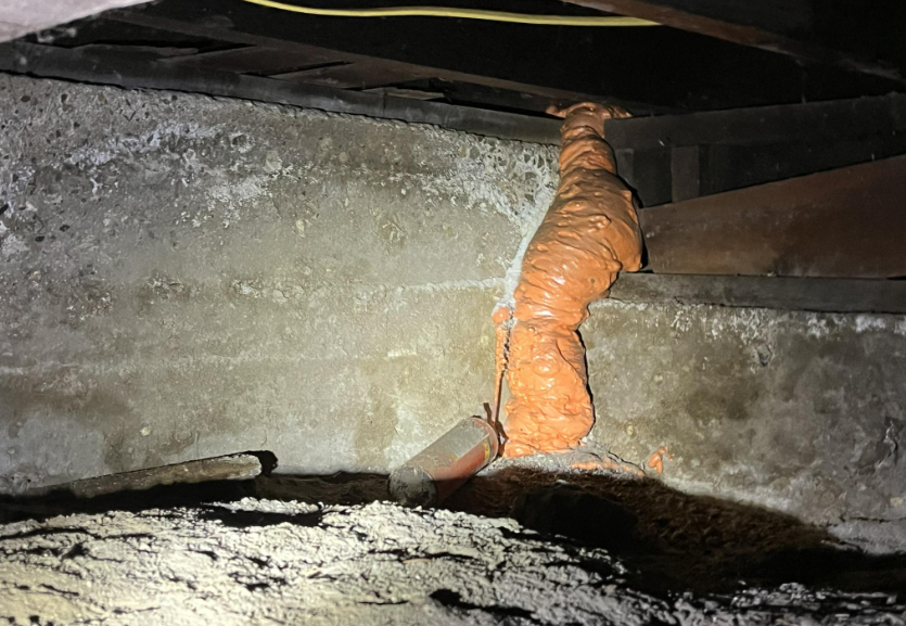 A dimly lit space with an exposed concrete wall, featuring an irregular and lumpy patch of orange foam insulation applied over a pipe. The foam extends along the wall and ceiling junction, with some loose debris visible on the dirt-covered floor below.