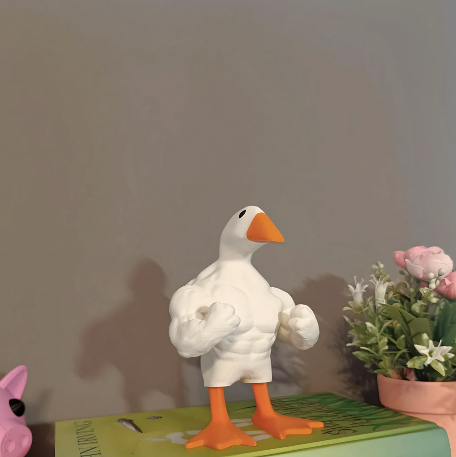 A figurine of a muscular duck with an orange beak and orange webbed feet is flexing its arms. It is placed on top of a green book near a pink flowerpot containing artificial flowers. In the background, there is a small portion of a pink object visible.